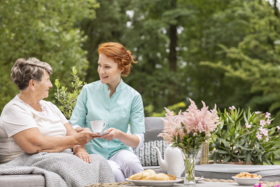 Happy nurse giving tea to elder woman while eating breakfast on the terrace